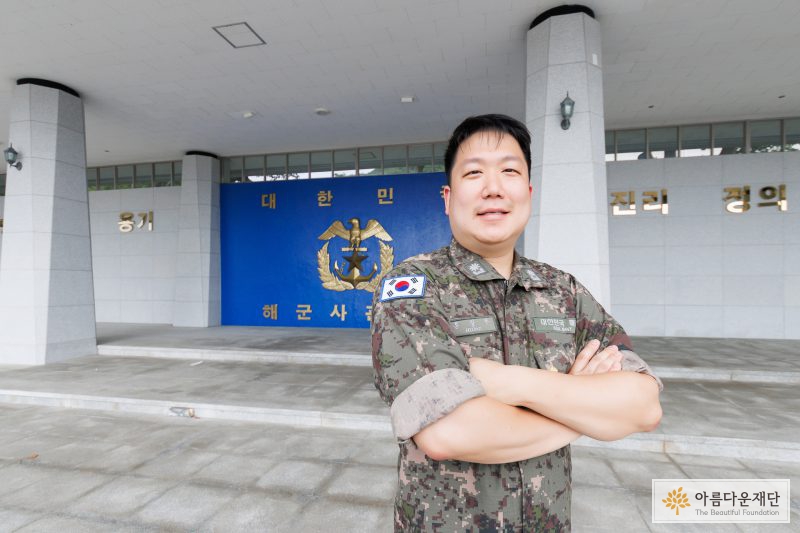 홍창우 기부자가 파란 배경에 금색으로 적힌 해군사관학교 마크가 있는 사열대 위에서 멋진 포즈를 취하고 있다. 홍창우 기부자는 밝게 웃고 있다.
