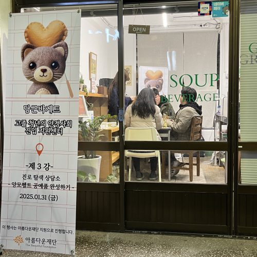 고졸청년의 안정사회 진입 지원센터라고 써 있는 배너 뒤로 카페 안에 앉이있는 사람들의 모습이 보인다. 진로탐색상담소 양모펠트 공예품 완성하기라고 배너 밑에 써 있다.