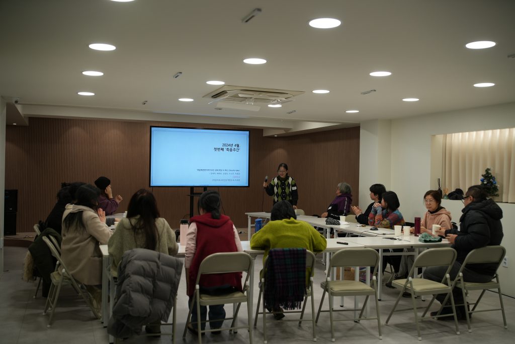 여러 명의 사람들이 책상에 둘러 앉아있고, 한 명이 화면 앞에서 무언가를 설명하고 있다. 