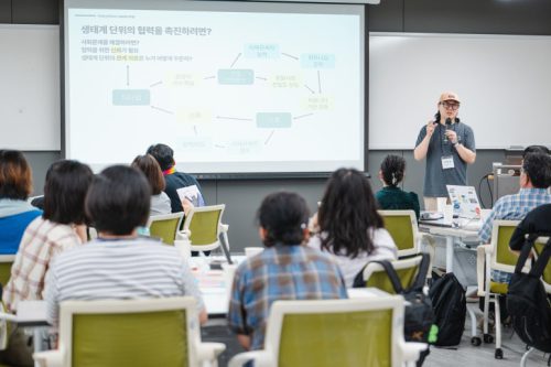 교육 강의자가 마이크를 잡고 말하고 있고, 참여자는 집중해서 듣고 있다.