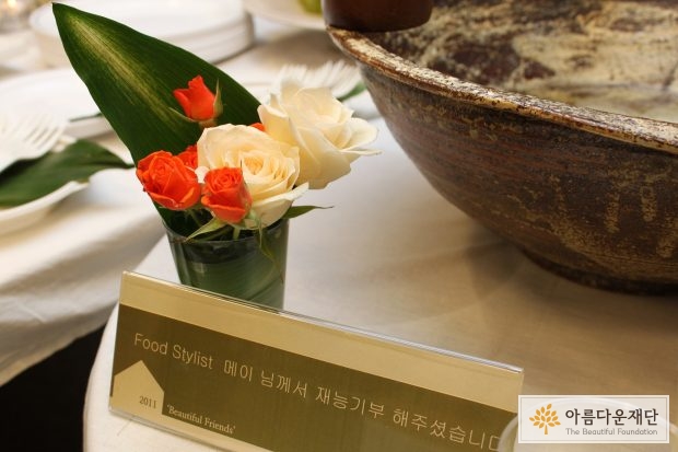 테이블위에 작은 꽃이 컵에 꽂혀있고 명패에 푸드스타일리스트 메이님의 재능기부가 적혀있다.